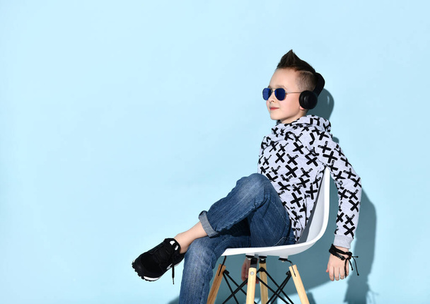 Teenage kid in headphones, sunglasses, blue jeans, hoodie, sneakers. Showing victory sign, sitting on white chair. Blue background - Фото, изображение