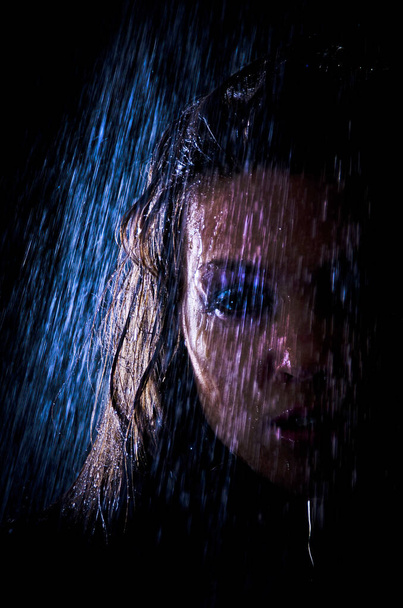 Conceptual view and scene of a young, sensual, beautiful girl, a woman who washes in the shower under a powerful jet of water in the backlight in an aqua studio on a dark background. - Valokuva, kuva
