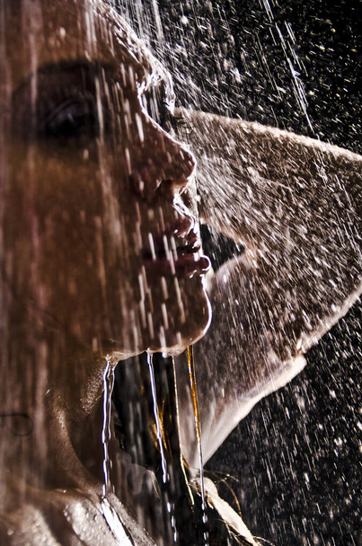 Conceptual view and scene of a young, sensual, beautiful girl, a woman who washes in the shower under a powerful jet of water in the backlight in an aqua studio on a dark background. - Foto, Bild