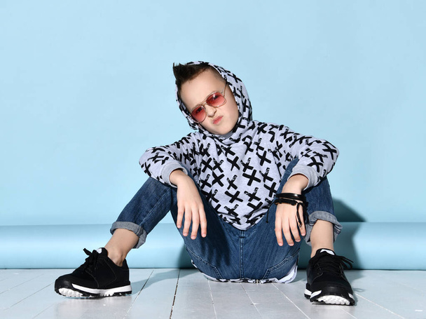 Teenager in sunglasses, blue jeans, hoodie, black bracelet and sneakers. Raised hand up, sitting on white floor. Blue background - Фото, изображение