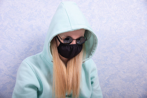 Egy fiatal nő fekete újrahasznosítható vírusvédő maszkban, fekete szemüvegben és kapucnis kabátban dolgozik otthon egy számítógépen. A távmunka fogalma otthon - Fotó, kép