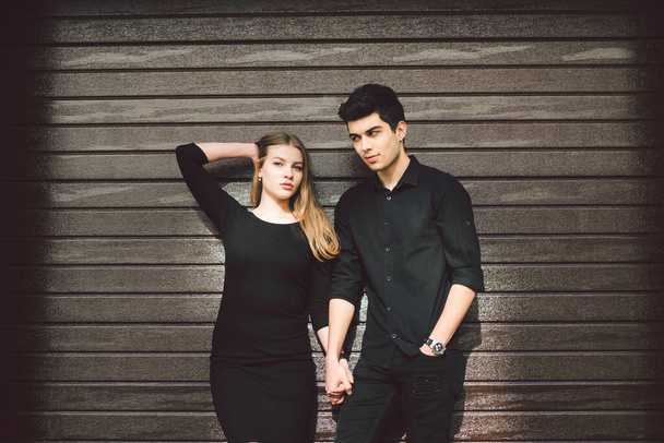 Portrait of attractive mixed race couple standing together against the background of a wooden wall. Dating, multiethnic wearing black clothes. Stylish young couple is spending time together outdoors. - Photo, Image
