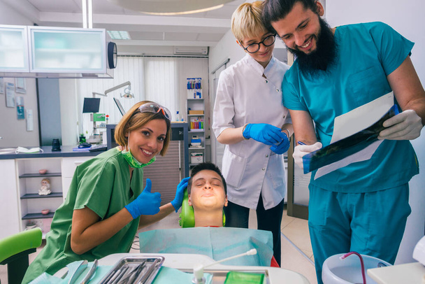 Porträt eines männlichen Zahnarztes, der einem jugendlichen Patienten ein Röntgenbild zeigt, während ihm seine lächelnden weiblichen Kollegen assistieren. - Foto, Bild