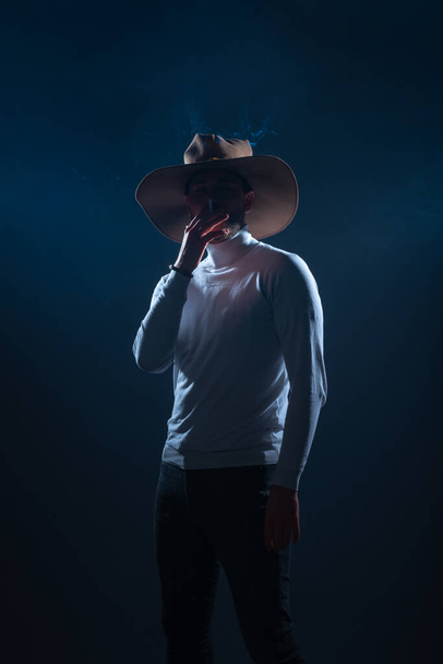 Portrait of a handsome young man with cowboy hat smoking cigarette in a studio on high contrast and dark background - Foto, imagen