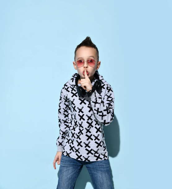 Teenage child with headphones on neck, in sunglasses, hoodie, black bracelet. He showing be quiet sign, posing on blue background - Photo, Image