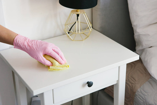 Καθαρισμός και απολύμανση σπιτιού: μια γυναίκα με γάντια καθαρίζει λευκά έπιπλα από τη σκόνη. - Φωτογραφία, εικόνα