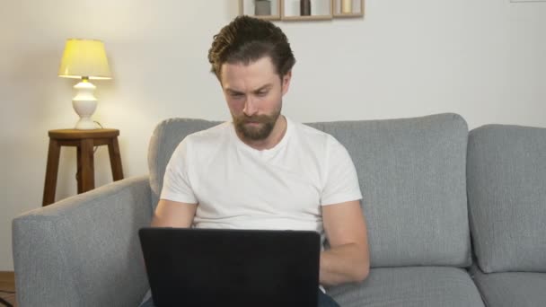 mladý muž sám sedí doma na gauči, na svém notebooku, hledá práci on-line, izolovaný od společnosti kvůli karanténě - Záběry, video