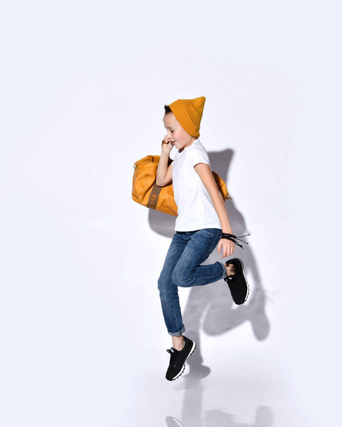 Teenage boy with orange travel bag, in hat, t-shirt, blue jeans, black sneakers. He jumping up, smiling, posing isolated on white - Фото, изображение