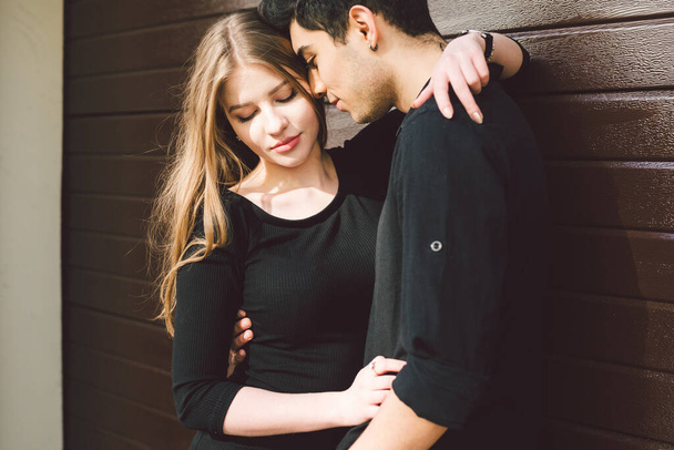 Portrait of attractive mixed race couple standing together against the background of a wooden wall. Dating, multiethnic wearing black clothes. Stylish young couple is spending time together outdoors. - Photo, Image