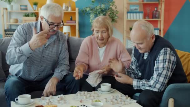 Joyful senior people enjoying lotto game indoors at home talking entertaining - Filmati, video