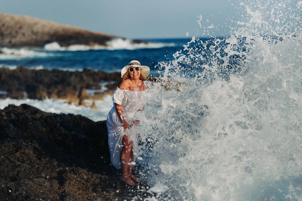 A beautiful girl in a white dress and a straw hat stands on the shore under the large oxen of the Atlantic Ocean. Big waves near her legs. - Photo, image