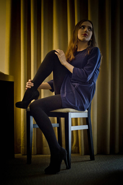 Conceptual portrait of a young, seductive, emotional, beautiful girl, a woman in a dress and tights, stockings and shoes sitting on a chair in a hotel room, in the interior. - Фото, изображение