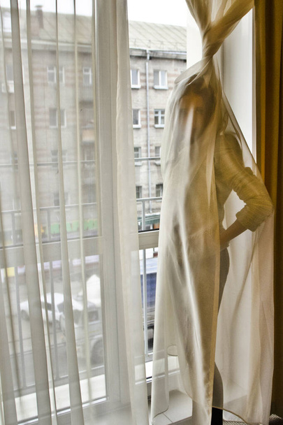 Conceptual portrait of a fashionable young, seductive, emotional, beautiful girl, a woman who wrapped herself in a curtain, tulle, in the interior of the apartments, hotel room - Zdjęcie, obraz