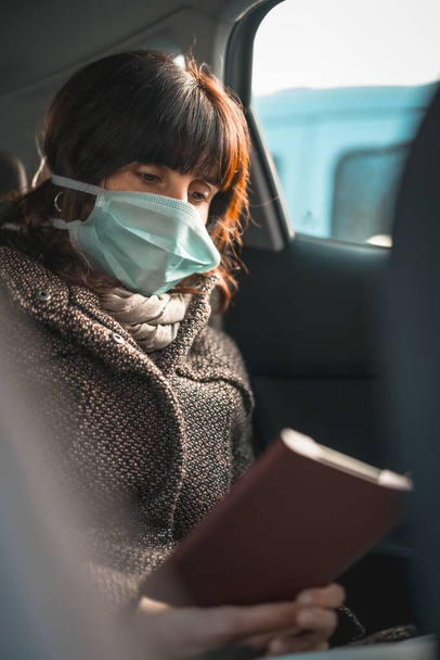 A young woman wearing a face mask sitting in the car and sadly reading an e-book - Fotografie, Obrázek