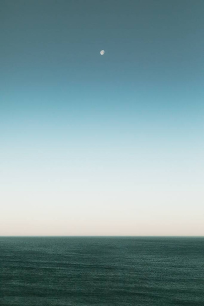 A vertical shot of a beautiful view of ocean under the moon in the blue sky - Fotó, kép