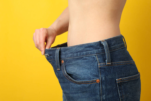 Chica tirando de sus pantalones vaqueros grandes y mostrando la pérdida de peso en el fondo amarillo
 - Foto, imagen