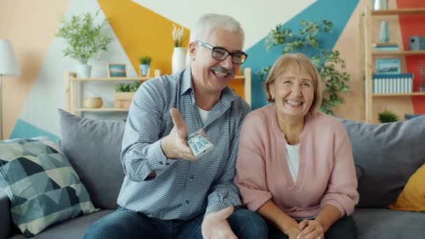 Portrait of happy old couple watching TV at home holding remote enjoying funny film - Video