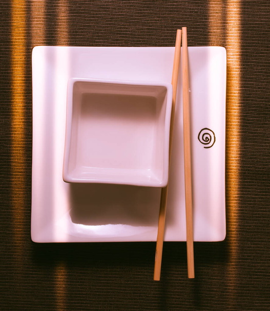 Square white plate and chopsticks in front of lighted window - Photo, Image