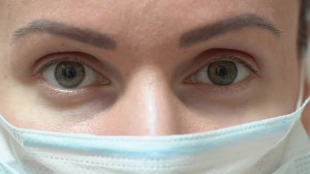 Dois olhos verdes de menina bonita olhando para a frente para a câmera em azul protetor máscara facial médica close-up. Retrato de uma jovem cirurgiã caucasiana. Quarentena durante a pandemia. Visão intensa
. - Filmagem, Vídeo