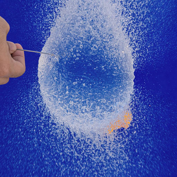 A vertical closeup of a person popping a water balloon with a needle against a blue background - Foto, imagen