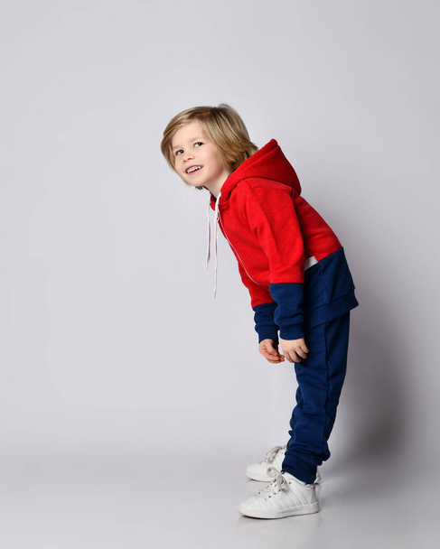 Happy smiling blond kid boy in blue and red hoodie and pants poses sideways bending over reaching his hands to toes - Foto, imagen