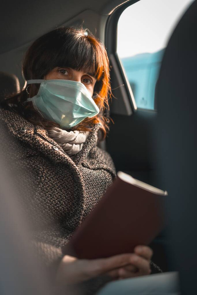 A young woman wearing a face mask sitting in the car and sadly reading an e-book - Foto, afbeelding
