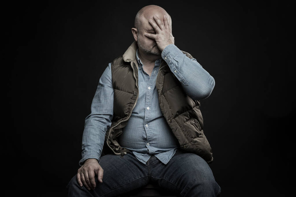 Ένας φαλακρός ενήλικας κρατάει το κεφάλι του στα χέρια του. Κατάθλιψη και οικονομικά προβλήματα σε μια κρίση. Ανεργία κατά τη διάρκεια της πανδημίας του κορωναϊού. Μαύρο φόντο. - Φωτογραφία, εικόνα
