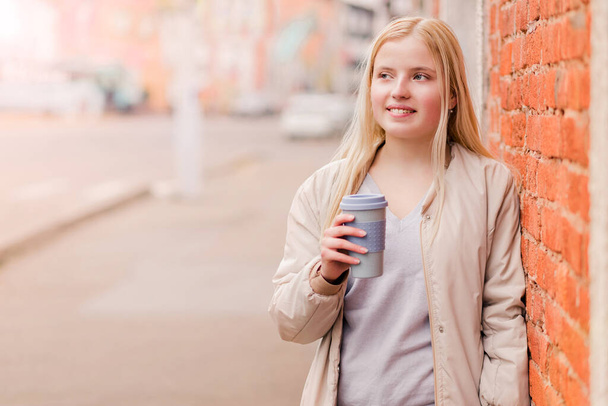 Portret van een jonge blanke vrouw met een kop koffie tegen een bakstenen muur. Stedelijk concept. - Foto, afbeelding
