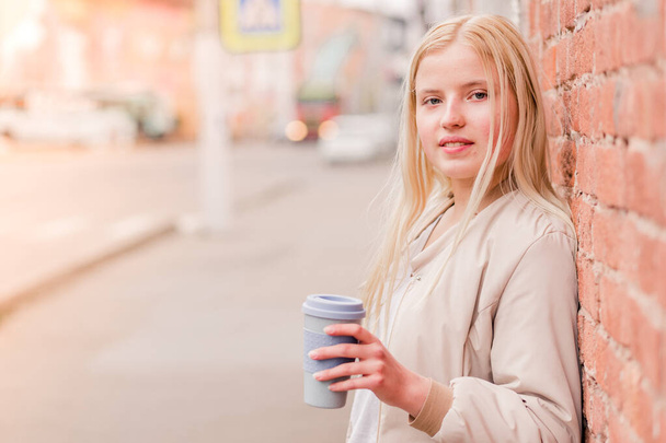 Egy fiatal fehér nő portréja, aki egy csésze kávét tart a téglafalnak. Városi koncepció. - Fotó, kép