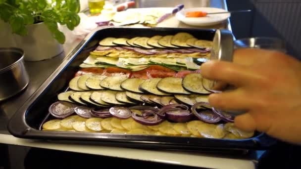 Przygotowanie mieszanych warzyw pokrojonych w cienkie plasterki, naoliwionych i umieszczonych na patelni do wypieku. Bakłażany, pomidory, ziemniaki, cukinie i czerwona cebula - Materiał filmowy, wideo