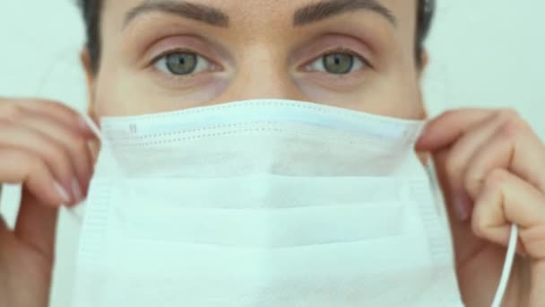 Gorgeous woman with green eyes nurse put on blue protection medical face mask on white background close up. Portrait of doctor during epidemic situation. COVID-19 or corona virus pandemic year. - Filmmaterial, Video