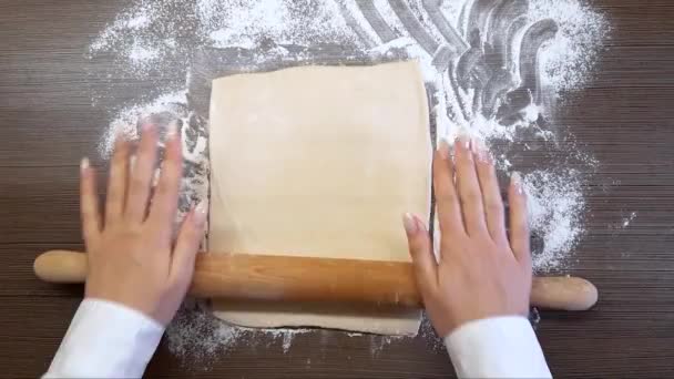 Kobiece ręce rozwałkowują ciasto drewnianym wałkiem do ciasta, na powierzchni stołu. Widok z góry - Materiał filmowy, wideo