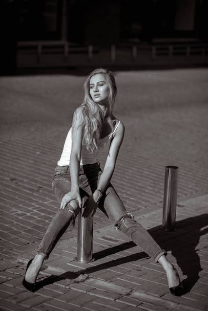 Sexy blonde woman wearing casual ripped jeans and t shirt, posing at the parking fence. Monochrome toning - Foto, imagen