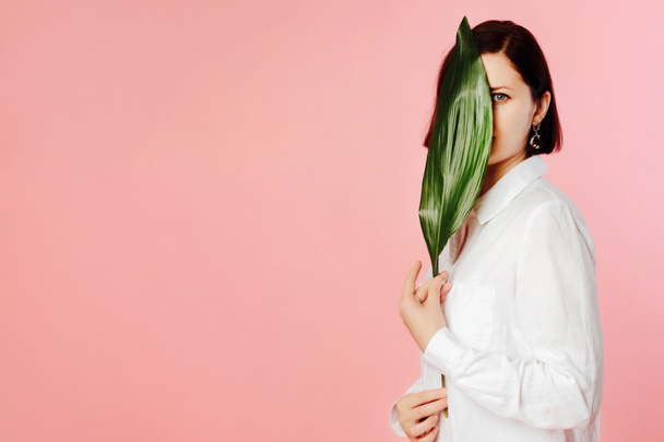 Ritratto di giovane donna che indossa camicia bianca, in piedi su sfondo rosa con foglia di palma verde. Ragazza guardando da dietro foglia di palma verde
. - Foto, immagini