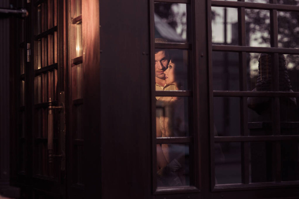 Młody mężczyzna obejmuje swoją kobietę od tyłu. Są za szklanymi drzwiami. W retro garniturach. Rekonstrukcja historyczna - Zdjęcie, obraz