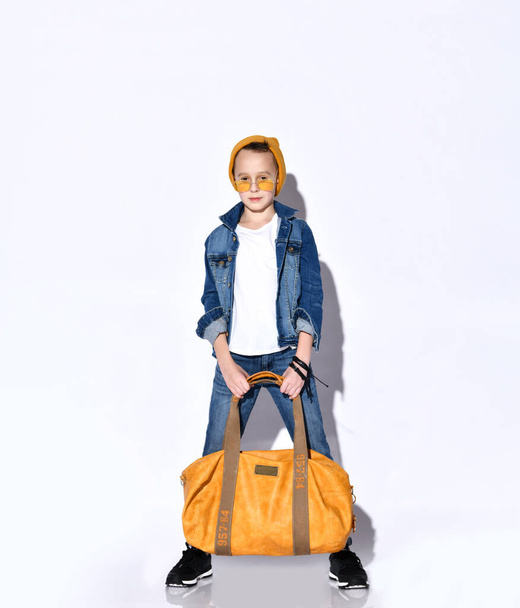 Teenage child with orange travel bag, in hat, sunglasses, t-shirt, blue denim suit, black sneakers. He posing isolated on white - Φωτογραφία, εικόνα