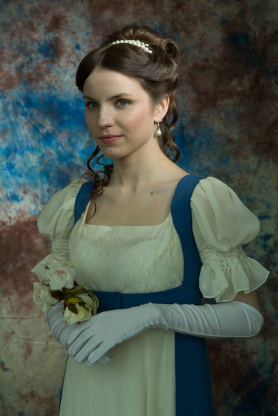 Una joven con un vestido largo de azul y blanco. Mira para otro lado. Sostiene flores blancas. Una mujer con un vestido retro. Reconstrucción histórica
 - Foto, imagen