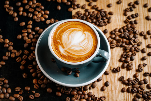 Tasse grise de café sur la table noire et brune parmi les grains de café torréfiés renversés. Cappuccino avec cofee art dans le café
. - Photo, image