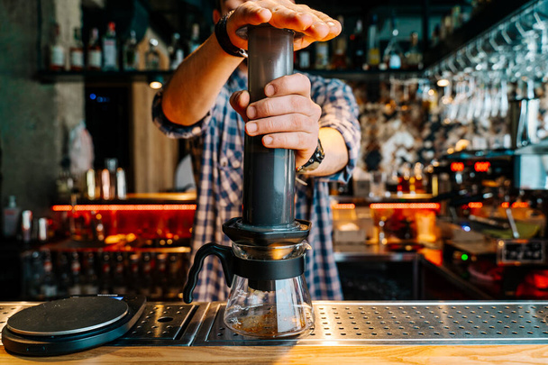 Alternative Coffee Brewing Method. Close-up of the hands of barista who is preparing filter coffee in an aeropress. Barista wears a checkered flannel shirt and works at the bar counter. - Photo, image