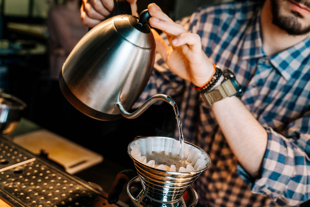 代替コーヒー醸造法。レストランでは、ケトルからコーヒーフィルターに水を注ぐバリスタの手のクローズアップ. - 写真・画像