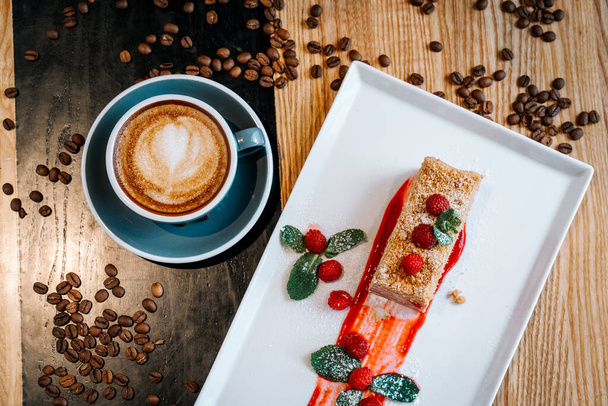 Cappuccino umění v šedém šálku s kusem napoleonského dortu s malinovým a mátovým listem mezi kávovými zrny na dřevěném stole v restauraci. Koncept milovníka kávy pro ranní snídani - Fotografie, Obrázek