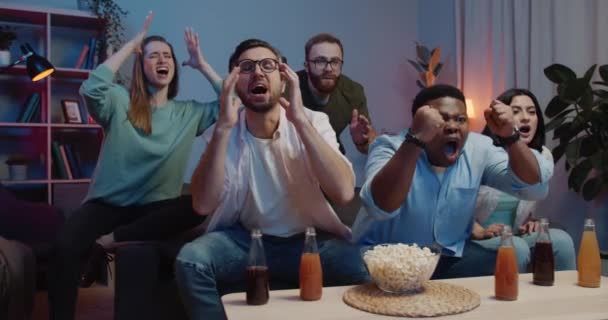 Multi ethnic group of men and women supporting team while watching sports on TV. Excited group of friends sitting on sofa in front of table with popcorn and drinks and getting upset. - Footage, Video