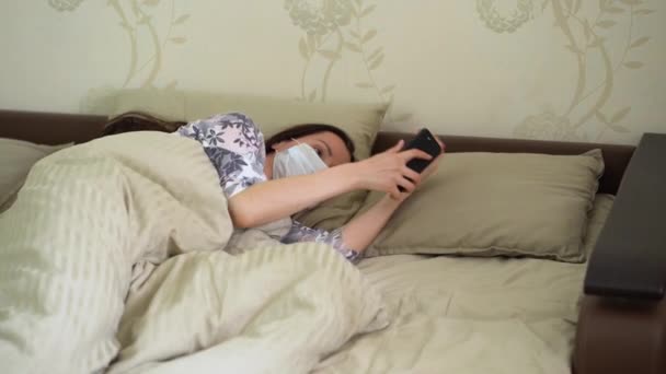 Young Caucasian girl in pajamas in protective medical mask sweet sleep under beige blanket in bed, wake up, turn off alarm ring on black phone, stretch, checks the social feed. Life in period pandemic - Footage, Video