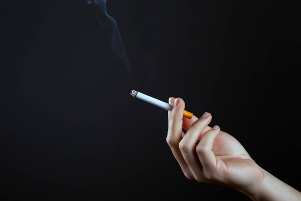 mano femenina sosteniendo un cigarrillo humeante en un espacio de copia de fondo oscuro. daño por fumar cigarrillos
 - Foto, imagen