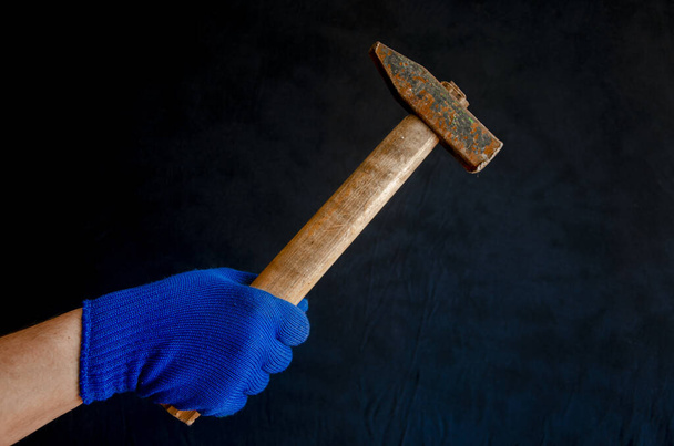 Ανδρικό χέρι σε μπλε γάντι με σφυρί στο χέρι σε σκούρο φόντο - Φωτογραφία, εικόνα