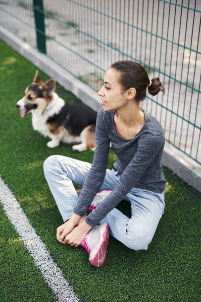 Young female owner sitting on grass with her cute pet Welsh Corgi dog outdoors, spending time together. Concept friendship with dog and human, dog walking - Фото, зображення