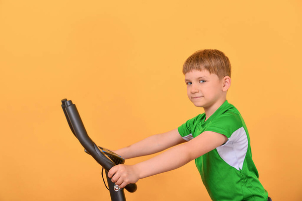 Το αγόρι πηγαίνει μέσα για σπορ σε ένα σταθερό ποδήλατο και κάνει προπόνηση καρδιο. - Φωτογραφία, εικόνα