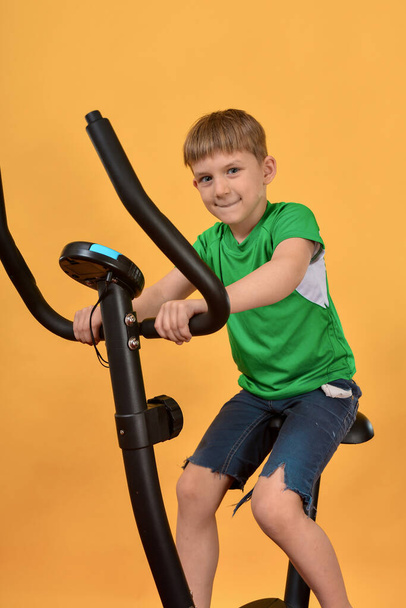 Мальчик занимается спортом, ребенок на стационарном велосипеде занимается кардио и смотрит в камеру
. - Фото, изображение