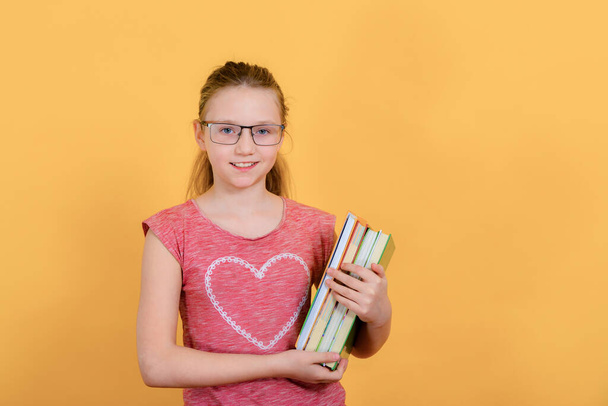 Ders kitapları olan neşeli bir genç kız sarı bir arka planda elinde bir kitapla duruyor.. - Fotoğraf, Görsel
