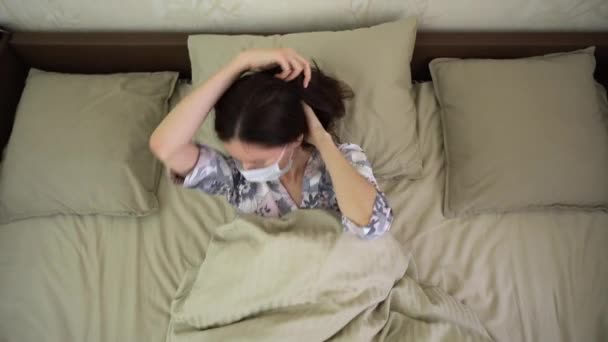 毛布の下に服を着た孤独な若い白人女性は、白い保護医療面マスクを枕の上に置く。病気の女の子のベッドで一番上のビューの真ん中で寝ることになる。住宅隔離の概念. - 映像、動画